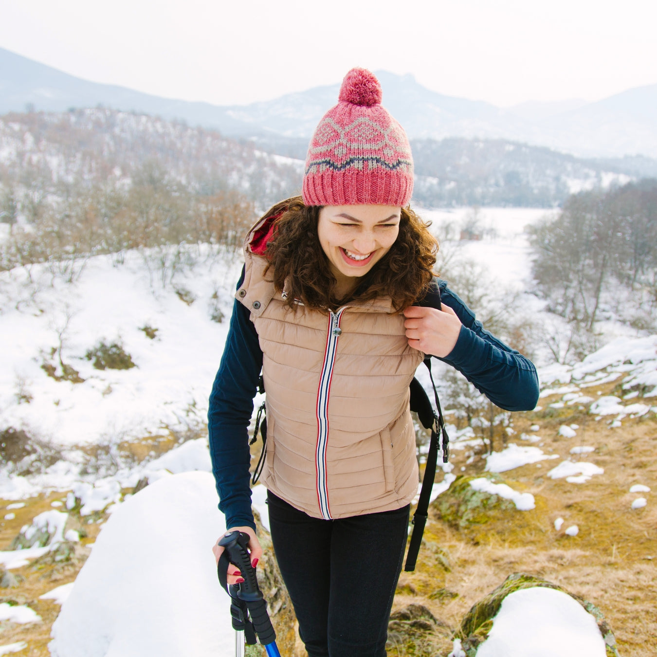 Woman hiking in a snowy landscape wearing a pink beanie and beige vest.