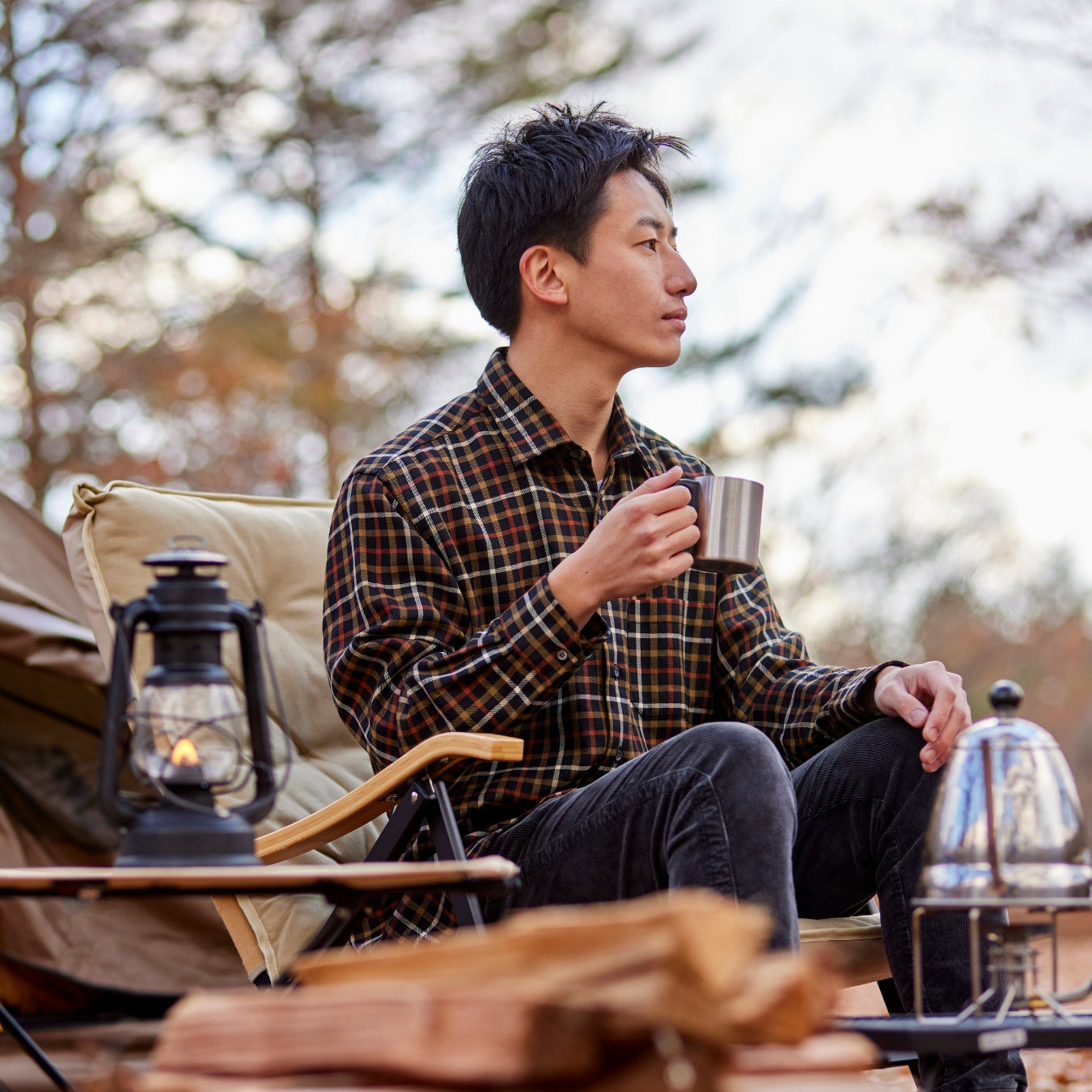Man sitting outdoors holding a mug, surrounded by camping gear with a blurred forest background